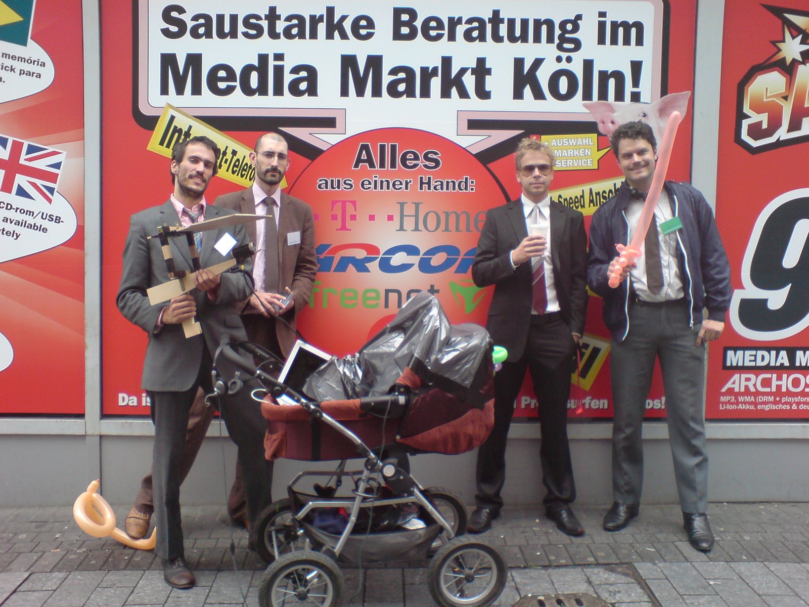 Four men in suits with signs and an equipment-rigged pram in front of Media Markt Cologne